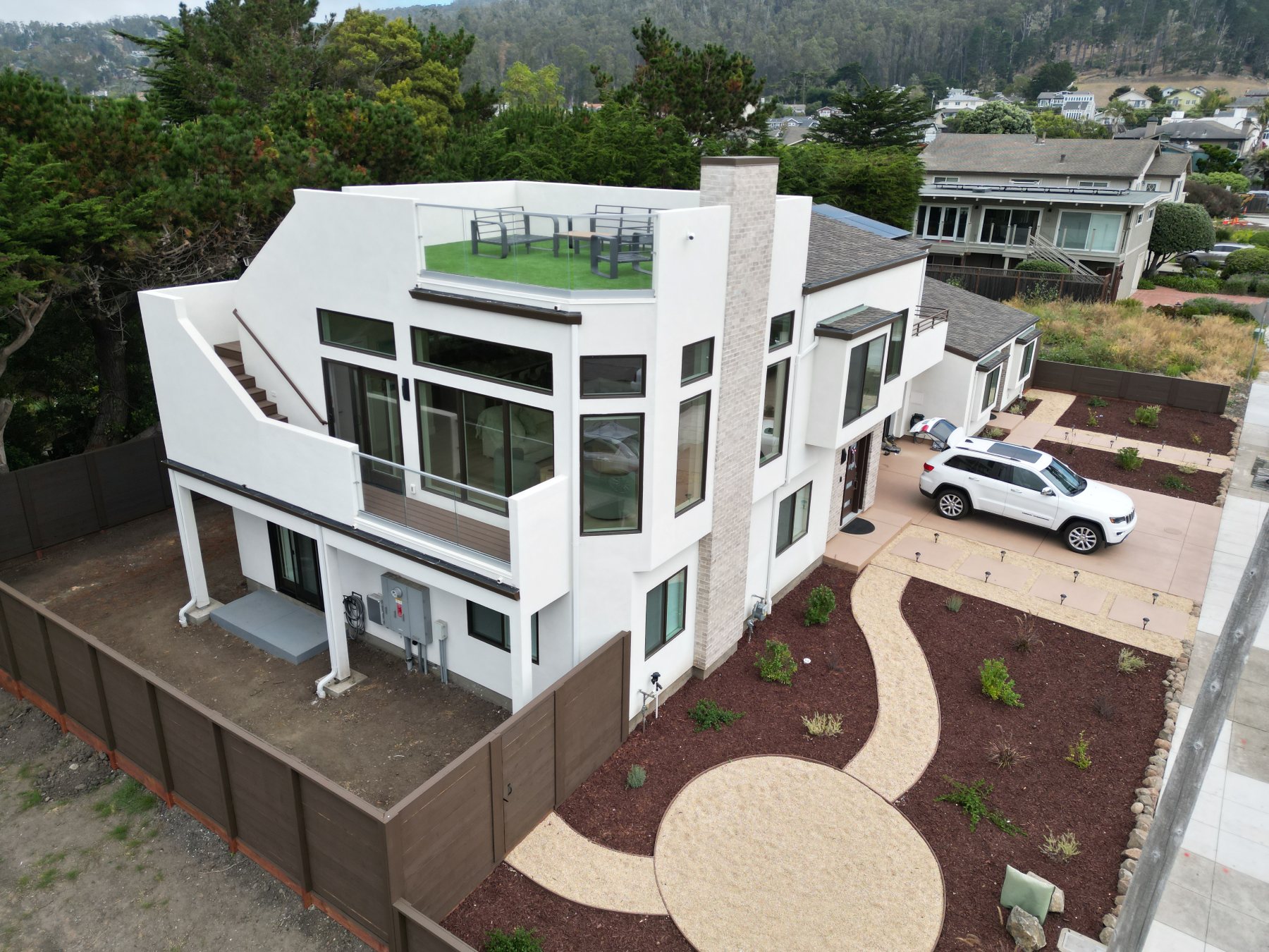 Aerial view from the rear, showing the rooftop deck, glass railings, and floor-to-ceiling windows facing the hillside.