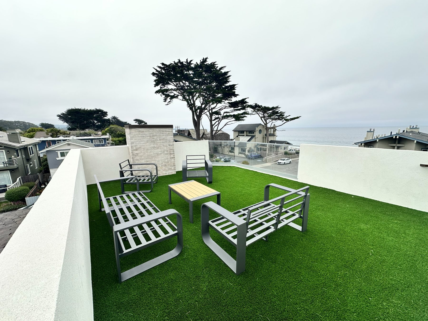 Rooftop deck with modular outdoor furniture and turf, framed by cypress trees with an ocean view beyond.