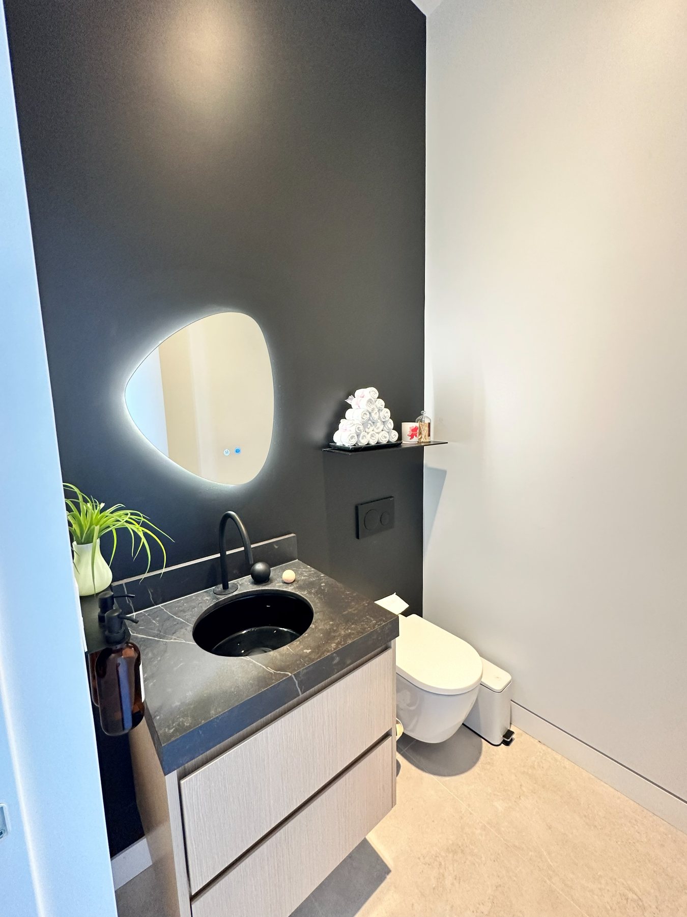 A powder room finished in black stone, with a sculptural backlit mirror.