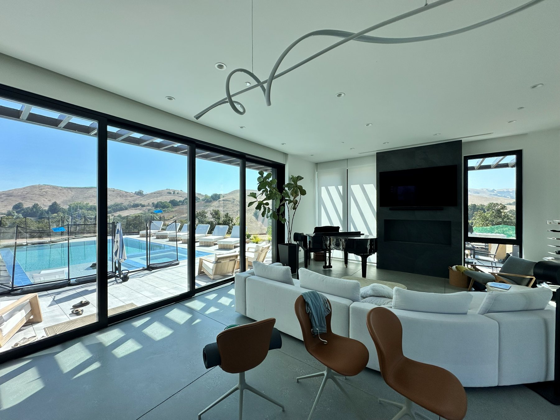 Living room with a grand piano and linear fireplace, facing floor-to-ceiling glass toward the pool.